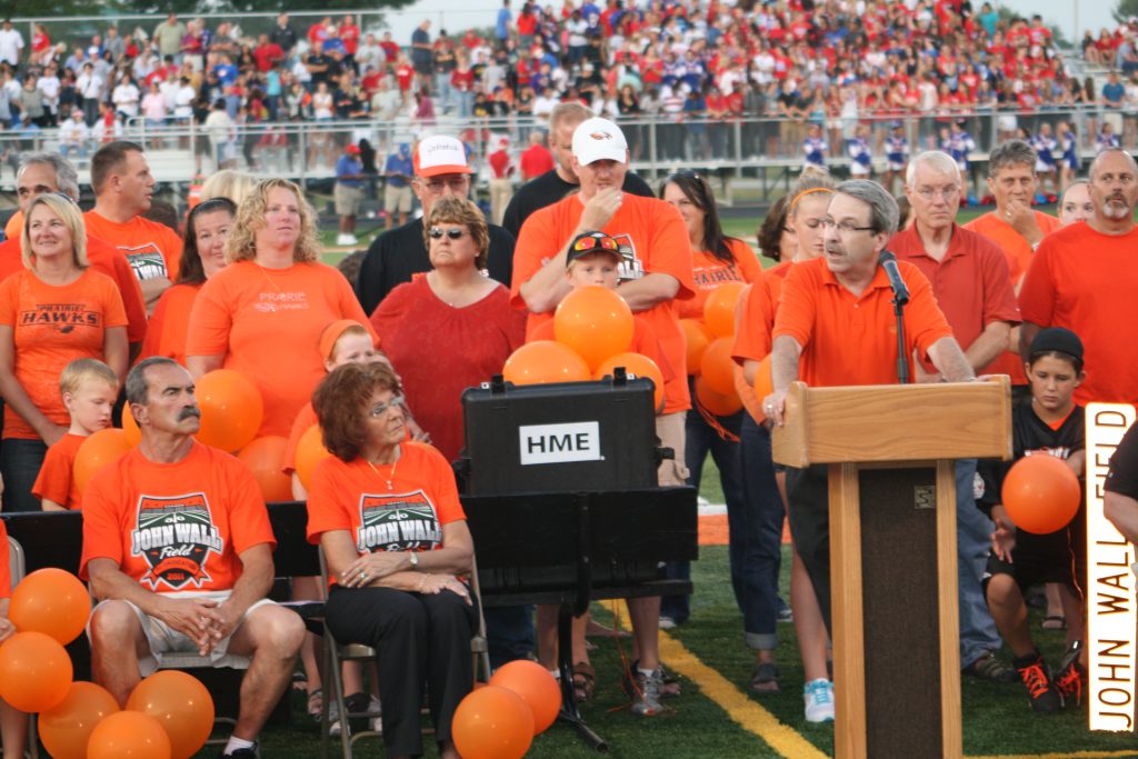 School Board President Randy Bauer standing behind a podium speaking to an audience at the John Wall rededication event.